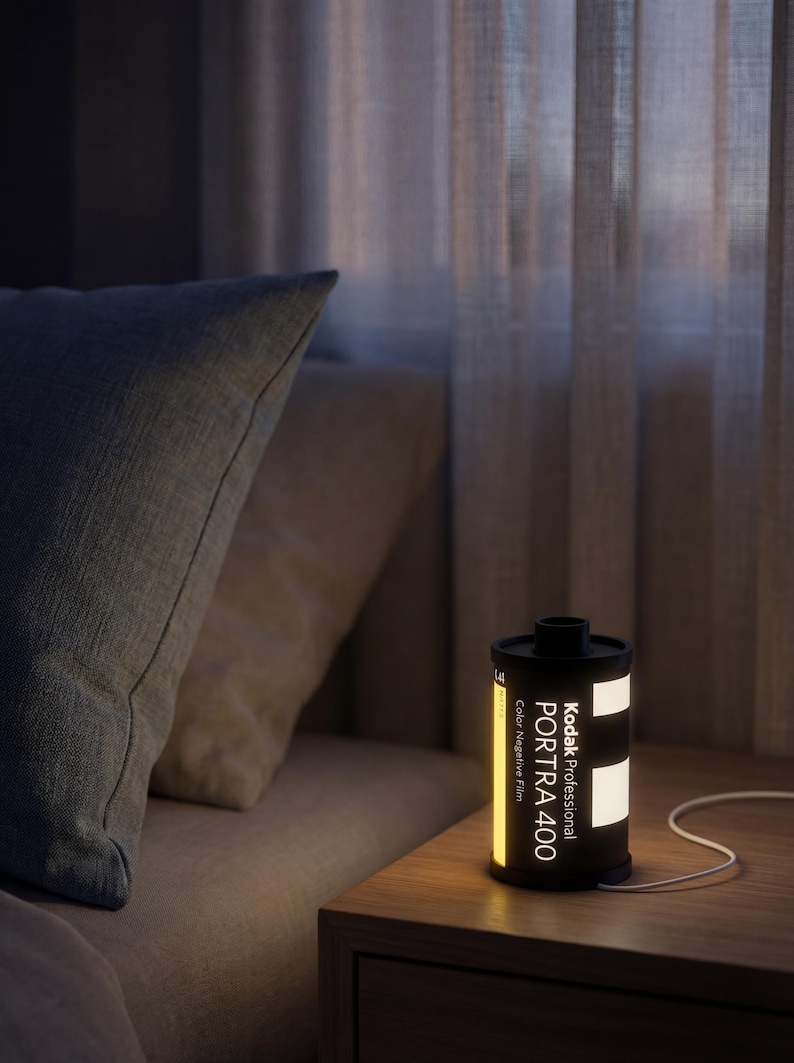 May include: A black and yellow Kodak Professional Portra 400 film canister lamp on a wooden bedside table. The lamp is illuminated, displaying the text "Kodak Professional Portra 400 Color Negative Film". A white cable extends from the lamp.