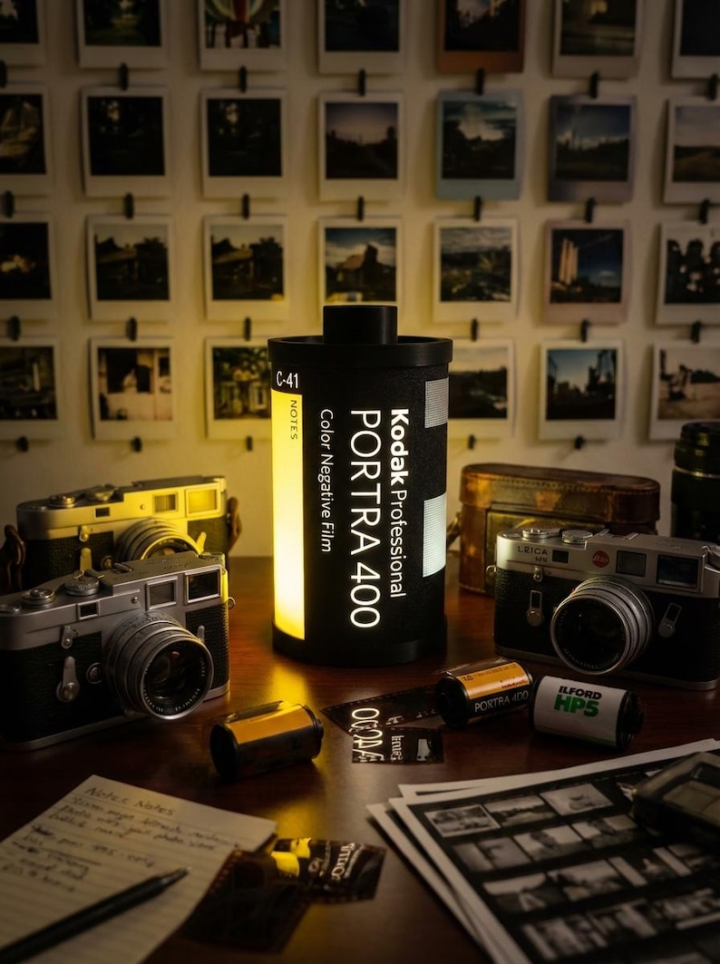 May include: A collection of vintage cameras, film rolls, and a Kodak PORTRA 400 film canister lamp. The scene includes a wall of framed photographs, creating a nostalgic atmosphere. The lamp is black with white text.