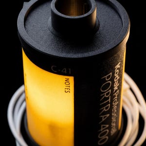 May include: A black and yellow Kodak Portra 400 film canister with white cord wrapped around it. The canister has the text "Kodak Professional PORTRA 400" and "NOTES" printed on it. The film is C-41.