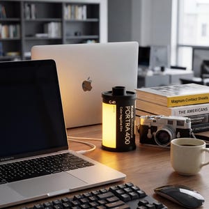 May include: A desk with a MacBook Pro, black keyboard, and mouse. A film canister lamp with "PORTRA 400" text is lit. A vintage camera, books, and a coffee cup are also present, creating a workspace scene.