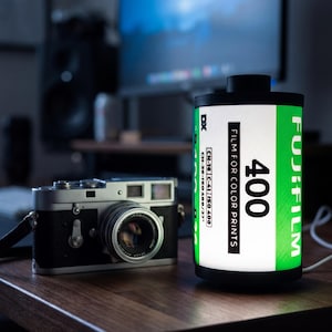 Peut inclure: Un appareil photo argentique noir et argenté à côté d'une lampe en forme de rouleau de film. La lampe porte une étiquette verte et blanche avec l'inscription "FUJIFILM 400 FILM FOR COLOR PRINTS".