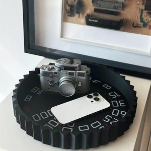 May include: A black circular tray with a gear-shaped edge and white numerical markings on its surface. A silver vintage camera and a white smartphone are placed on top of the tray. The tray features a series of numbers printed in a circular pattern.