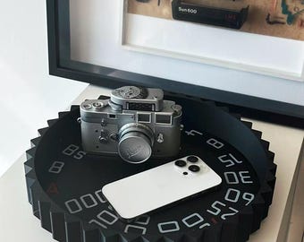 May include: A black circular tray with a gear-shaped edge and white numerical markings on its surface. A silver vintage camera and a white smartphone are placed on top of the tray. The tray features a series of numbers printed in a circular pattern.