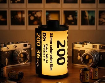May include: A film canister-shaped lamp, with the number "200" and "36 exp." printed on it. The lamp is lit, and the text "35mm color print film" is visible. Classic cameras and film rolls are arranged on a wooden surface.