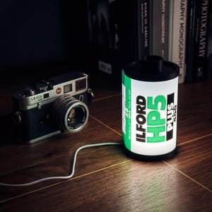 Peut inclure: Un appareil photo Leica M6 noir et argenté est posé à côté d'une lampe en forme de bobine de film éclairée. La lampe affiche "ILFORD HP5 PLUS 400" en vert et noir. Un câble blanc relie la lampe.