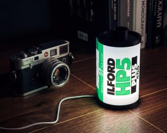 Peut inclure: Un appareil photo Leica M6 noir et argenté est posé à côté d'une lampe en forme de bobine de film éclairée. La lampe affiche "ILFORD HP5 PLUS 400" en vert et noir. Un câble blanc relie la lampe.