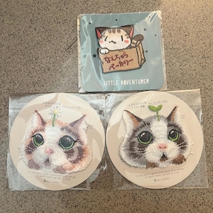 May include: Three embroidered cat patches in clear plastic packaging. One square patch shows a cat in a cardboard box with Japanese text. Two circular patches feature cat faces with a small flower or sprout on their heads and English text.