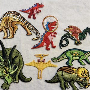Peut inclure: Un ensemble de huit écussons brodés représentant des dinosaures de différentes couleurs et espèces, incluant un grand tricératops vert, un ptérodactyle jaune avec un nœud papillon, un dinosaure rouge dans un casque spatial et d'autres dinosaures.