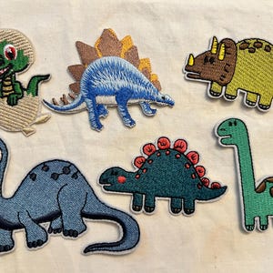 Puede incluir: Una colección de parches de dinosaurios bordados en varios colores. Los parches incluyen un dinosaurio verde saliendo de un huevo, un estegosaurio azul, un triceratops amarillo y marrón, un brontosaurio azul, un estegosaurio verde y rojo y un brontosaurio verde.