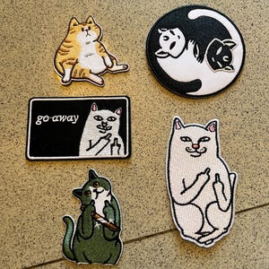 May include: Five embroidered patches featuring cats: a seated orange tabby, a circular black and white yin-yang cat design, a rectangular patch with a white cat and the text "go away", a green cat holding a knife, and a white cat gesturing with a middle finger.