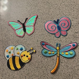 May include: Four embroidered patches featuring a teal butterfly with pink borders, a pink butterfly with blue swirls, a yellow and black striped bee with polka-dot wings, and a blue and orange dragonfly with circular patterns on its wings.