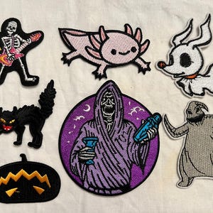 May include: A collection of embroidered patches featuring Halloween-themed designs. Includes a skeleton playing a guitar, a pink axolotl, a ghost dog, a black cat, a jack-o'-lantern, a grim reaper, and a burlap-textured character.