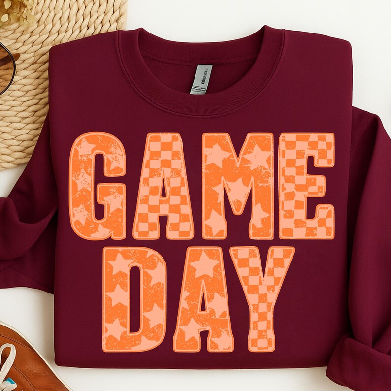 May include: Burgundy sweatshirt with the words "GAME DAY" in large orange letters. The letters have a combination of star and checkerboard patterns. The sweatshirt is folded, with a glimpse of a woven basket in the background.