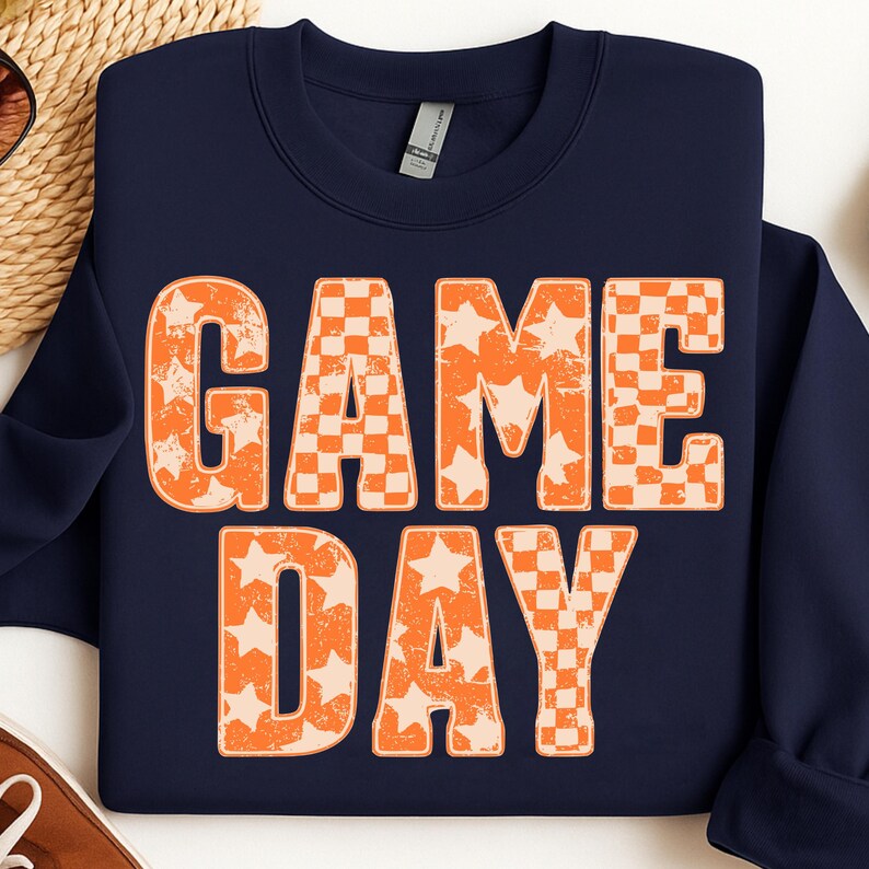 May include: Navy blue sweatshirt with the words "GAME DAY" in large orange letters. The letters are a combination of star and checkerboard patterns. The sweatshirt is folded and appears to be made of a soft material.