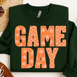 May include: Dark green sweatshirt with the words "GAME DAY" in large orange letters. The letters have a distressed look, with a mix of star and checkerboard patterns. A casual, sporty design.