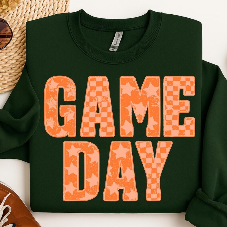 May include: Dark green sweatshirt with the words "GAME DAY" in large orange letters. The letters have a distressed look, with a mix of star and checkerboard patterns. A casual, sporty design.
