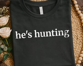 May include: Dark gray t-shirt with the text "he's hunting" in white, distressed font. The shirt is folded and displayed on a woven surface, with floral and beaded accents.