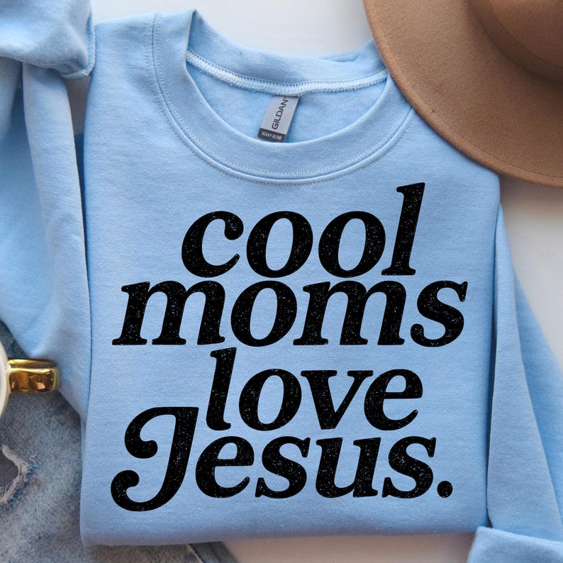 K&ouml;nnte beinhalten: Hellblaues Sweatshirt mit dem Text "cool moms love Jesus" in fetter, schwarzer Schrift. Das Sweatshirt ist gefaltet, im Hintergrund ist ein beiger Hut zu sehen. Das Gildan-Markenschild ist sichtbar.