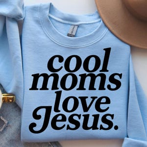 K&ouml;nnte beinhalten: Hellblaues Sweatshirt mit dem Text "cool moms love Jesus" in fetten schwarzen Buchstaben. Das Sweatshirt ist gefaltet, im Hintergrund ist ein brauner Hut zu sehen.