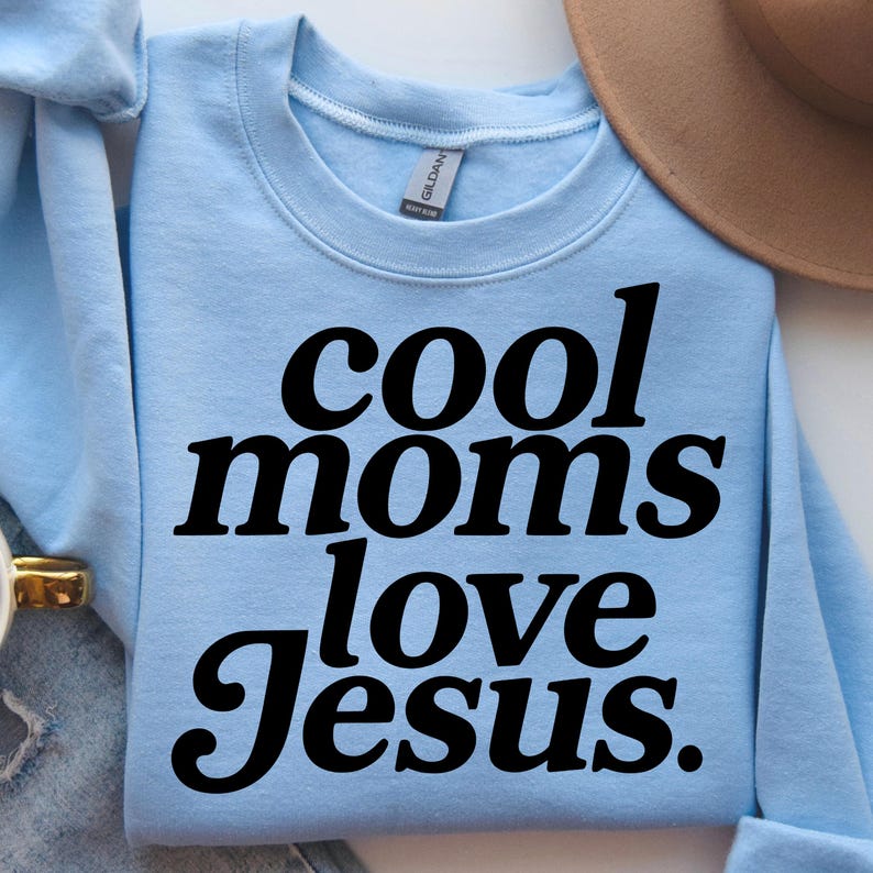 K&ouml;nnte beinhalten: Hellblaues Sweatshirt mit dem Text "cool moms love Jesus" in fetten schwarzen Buchstaben. Das Sweatshirt ist gefaltet, im Hintergrund ist ein brauner Hut zu sehen.