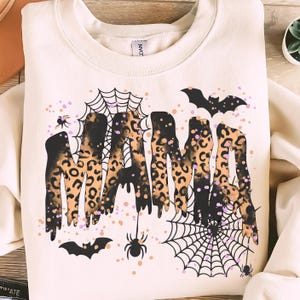 May include: Cream-coloured sweatshirt featuring the word "MAMA" in a leopard print design, with spiderwebs, bats, and spiders. The design has black, orange, and purple accents.