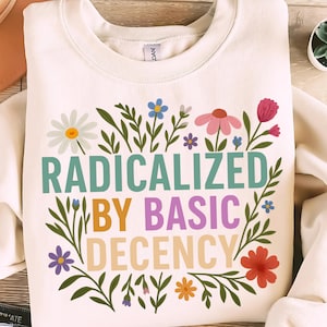 May include: Cream-colored sweatshirt with the text "RADICALIZED BY BASIC DECENCY" in teal, orange, purple, and white. The text is surrounded by a floral design with daisies, pink, blue, and orange flowers and green leaves.