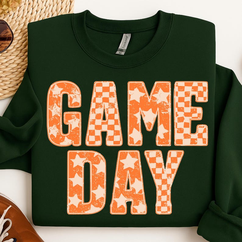 May include: Dark green sweatshirt with the words "GAME DAY" in large, distressed orange letters. The letters have a checkerboard and star pattern. The sweatshirt is folded on a white surface.