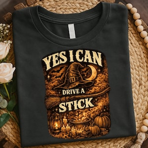May include: A black t-shirt featuring a graphic print with the text YES I CAN DRIVE A STICK. The design includes a witch hat, a broomstick, a crescent moon, bats, a lantern, candles, a cauldron, and pumpkins in shades of orange and brown.