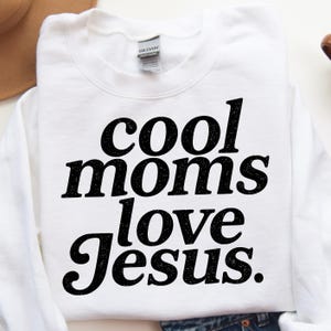 K&ouml;nnte beinhalten: Wei&szlig;es Sweatshirt mit dem Text "cool moms love Jesus" in fetten, schwarzen Buchstaben. Das Sweatshirt ist von der Marke Gildan. Der Text ist mittig auf der Vorderseite des Kleidungsst&uuml;cks platziert.