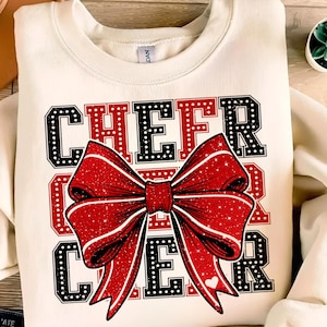May include: A white sweatshirt with the words "Cheer Cheer" in black and red letters with a red and white glitter bow in the centre.