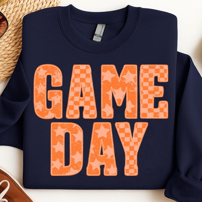 May include: Navy blue sweatshirt with the words "GAME DAY" in orange. The letters have a distressed look, with some featuring a star pattern and others a checkerboard design. A casual, sporty style.