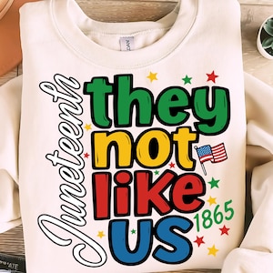 Peut inclure: Sweat-shirt crème avec le mot "Juneteenth" en écriture cursive. L'expression "they not like us" est en vert, jaune, rouge et bleu, avec un drapeau américain et l'année 1865.