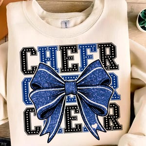 May include: Cream-colored sweatshirt with the word "CHEER" in black block letters outlined in white dots and blue letters. A large blue glitter bow is centered on the sweatshirt. A small white heart is on the bow.