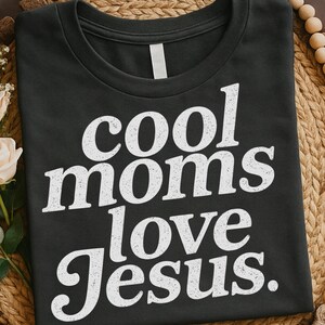 K&ouml;nnte beinhalten: Dunkelgraues T-Shirt mit dem wei&szlig;en Text "cool moms love Jesus" in fetter Serifen-Schrift. Das T-Shirt hat einen Rundhalsausschnitt und kurze &Auml;rmel. Der Text ist mittig auf der Vorderseite platziert.