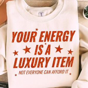 May include: A cream-coloured sweatshirt featuring the phrase "YOUR ENERGY IS A LUXURY ITEM" in glittery orange lettering, with star motifs. The text "NOT EVERYONE CAN AFFORD IT" is printed below. A casual, statement piece.