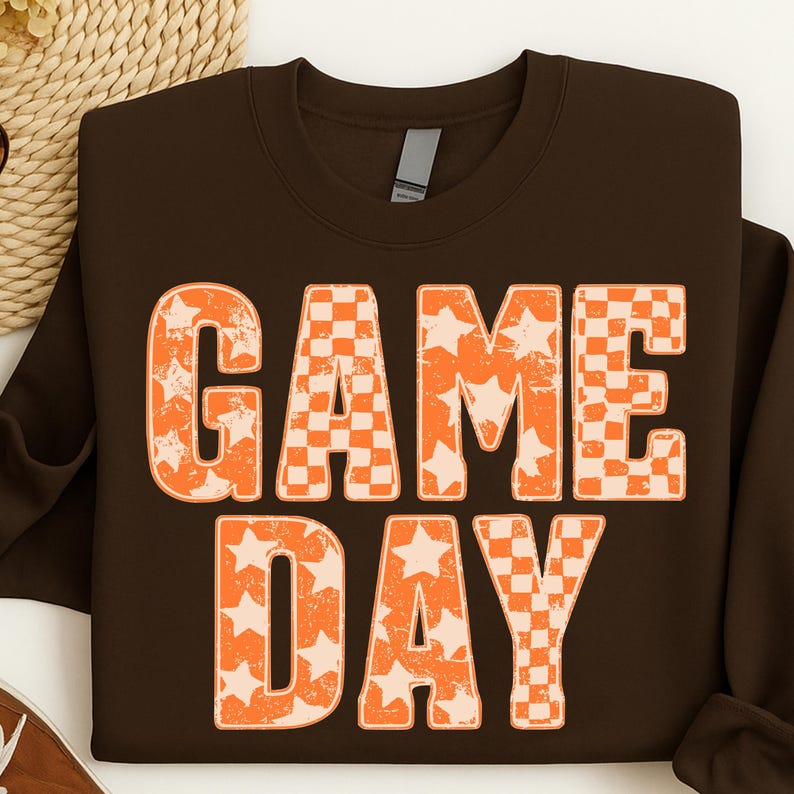 May include: A brown sweatshirt with the words "GAME DAY" in large, orange, distressed lettering. The letters are filled with a checkerboard pattern and stars. The sweatshirt has a crew neck and long sleeves.