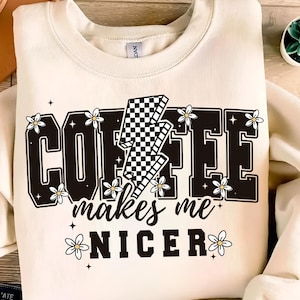 Pode incluir: Uma camisola de cor creme com um design gráfico preto e branco que diz "Coffee makes me nicer" com um raio xadrez e margaridas brancas.