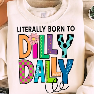 May include: Cream-colored sweatshirt with the text "LITERALLY BORN TO DILLY DALLY" in colorful, playful lettering. The word "DILLY" features a daisy, and the letters are in orange, green, purple, and teal. The word "DALLY" is in green, teal, orange, and blue.