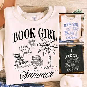 Op de afbeelding: Een crèmekleurig sweatshirt met zwarte zeefdruktekst die BOOK GIRL Summer leest. De afbeelding bevat een zon, een palmboom, een stapel boeken en een strandstoel met een parasol. Twee kleinere inzetfoto's tonen hetzelfde ontwerp.