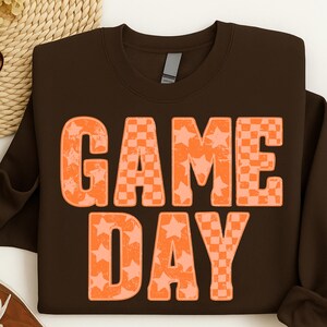 May include: A brown sweatshirt with the words "GAME DAY" in large orange letters. The letters are filled with a combination of star and checkerboard patterns. The sweatshirt has a crew neck and long sleeves.