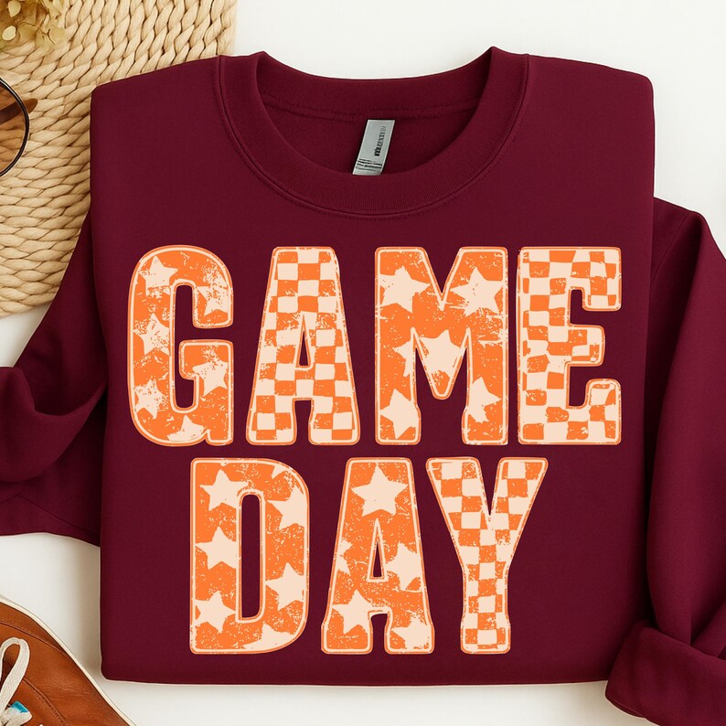 May include: A maroon sweatshirt with the words "GAME DAY" in large, orange letters. The letters have a mix of star and checkerboard patterns. The sweatshirt is made of a soft material.
