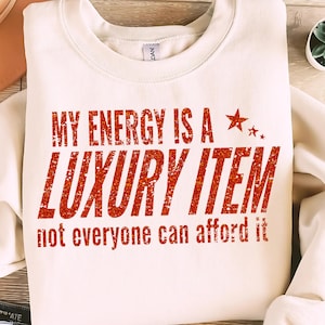 May include: A cream-coloured sweatshirt featuring the text "MY ENERGY IS A LUXURY ITEM not everyone can afford it" in red glitter. The design includes a star graphic.