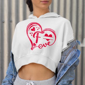 May include: White cropped hoodie with a red heart design. The heart has a large letter "F" inside and the word "love" written below. There is a small space inside the heart for a name.