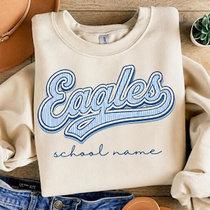 May include: A cream-colored crewneck sweatshirt featuring the word "Eagles" in a blue and white striped font with a blue outline, and the text "school name" in a simple blue script font centered below the main graphic on the chest.