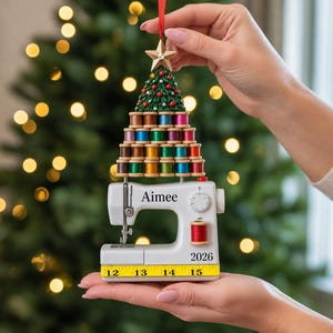 May include: A Christmas tree ornament featuring a white sewing machine with the name Aimee and the year 2026 printed on it. Above the machine is a pyramid of colourful thread spools topped with a green tree and a gold star, hanging by a red ribbon.