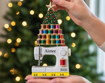 May include: A Christmas tree ornament featuring a white sewing machine with the name Aimee and the year 2026 printed on it. Above the machine is a pyramid of colorful thread spools topped with a green tree and a gold star, hanging by a red ribbon.