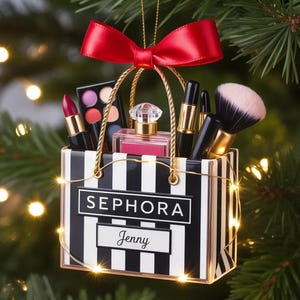 May include: A Sephora shopping bag ornament with black and white vertical stripes, featuring the name Jenny. The bag contains a perfume bottle, lipstick, eyeshadow palette, and makeup brush, topped with a red satin ribbon and gold cord.