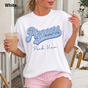 May include: A white short-sleeved t-shirt featuring the blue text Pioneers in a stylized script font with a striped pattern, followed by the text Peak View in a simple blue script font. The shirt is paired with light pink and white striped shorts.