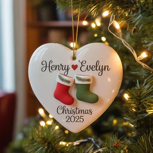 May include: White heart-shaped Christmas ornament with the names "Henry" and "Evelyn" and the year "2025". Features red and green stocking accents. The ornament is hanging on a Christmas tree.