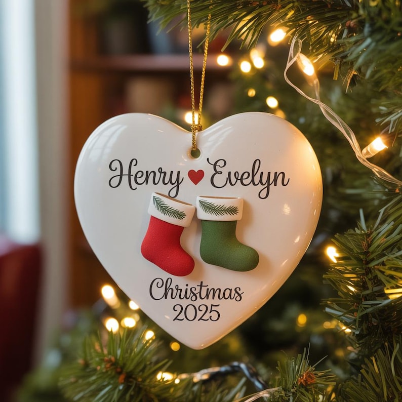 May include: White heart-shaped Christmas ornament with the names "Henry" and "Evelyn" and the year "2025". Features red and green stocking accents. The ornament is hanging on a Christmas tree.